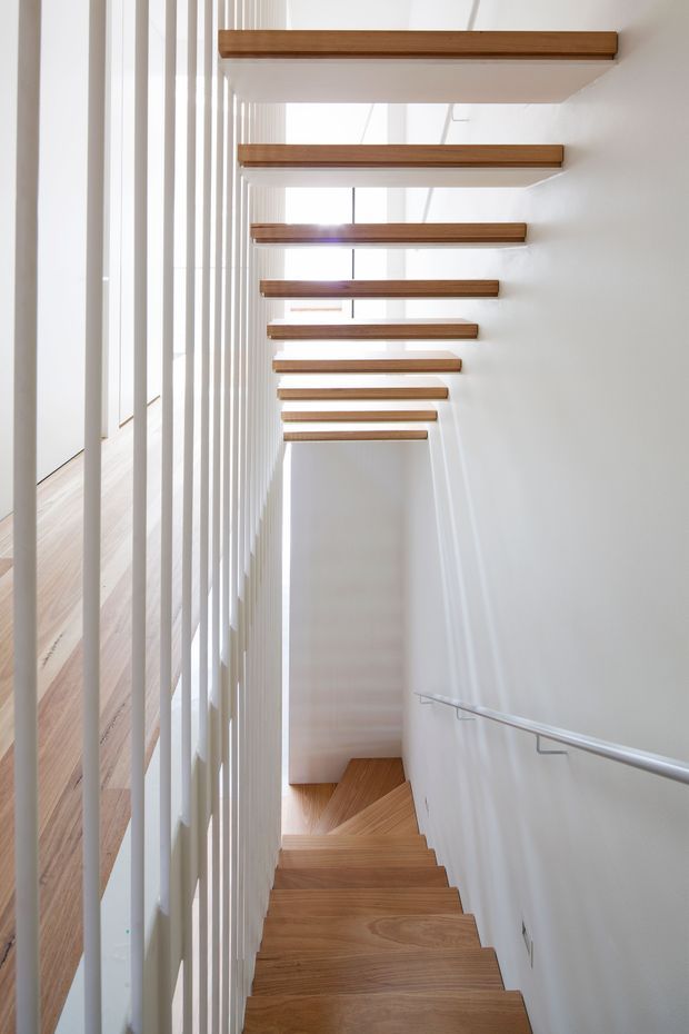 Bright contemporary stairway with warm timber treads and matching horizontal overhead beams, enclosed by white walls and a vertical white slat balustrade. A slim metal handrail runs down the right wall, and natural light filters through the stairwell.
