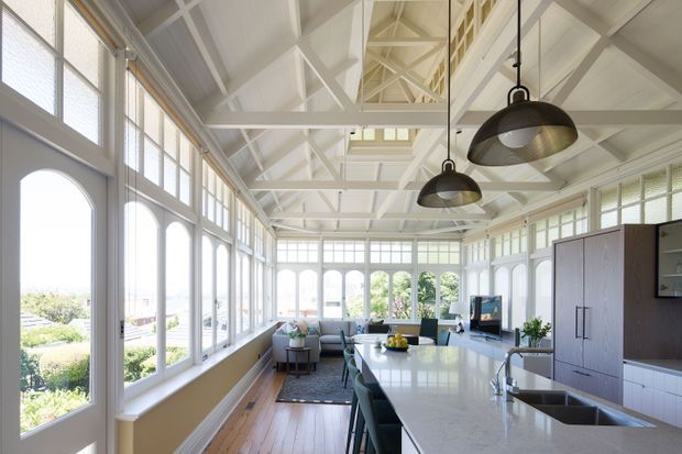 Bright, elongated open-plan kitchen and living space inside a highly glazed, pavilion-like room. White-painted structural trusses and wall panelling create a light architectural envelope, while large arched and rectangular windows overlook landscaped surroundings. A large stone island with integrated sink and dark stools anchors the kitchen, with a dining table, sofa and television area beyond. Black pendant lights provide contrast against the white ceiling.