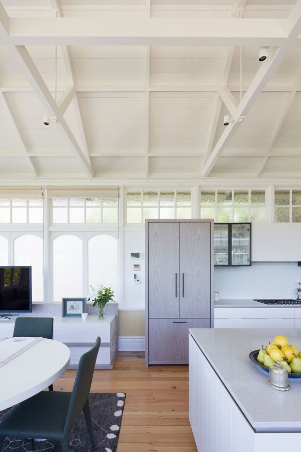 Bright open-plan kitchen and dining area with a white-painted high ceiling featuring exposed beams and trusses. The room has extensive textured and frosted glazing, white cabinetry, a pale stone island benchtop, white subway-tile splashback, timber flooring, and a tall grey timber pantry cabinet. A round dining table, dark upholstered chairs, and a small living area are also visible.