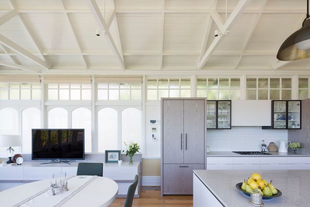 Bright open-plan kitchen and dining area with a large central island, white cabinetry, pale stone benchtops, timber flooring, and a freestanding-style integrated refrigerator. The room features extensive white-framed glazing with arched lower panels, exposed white-painted structural beams, a high ceiling, pendant lighting, and an adjacent dining table and living area with television.