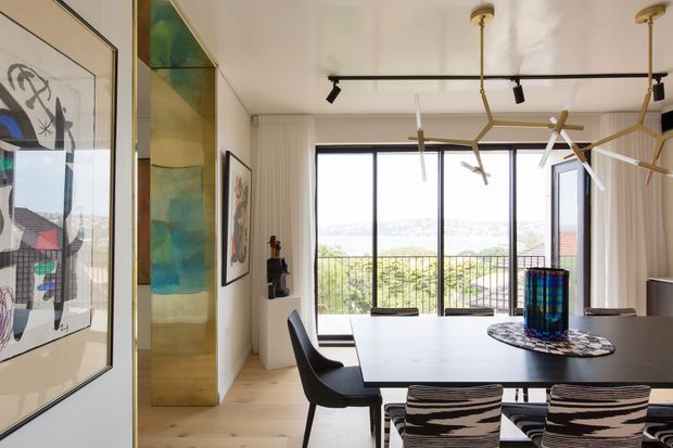 A bright, contemporary dining room with a long dark dining table and sculptural black chairs, opening onto a balcony through full-height windows. The room features pale timber flooring, textured light walls, large framed artwork, a colourful translucent feature panel, and striking branch-like pendant lighting.