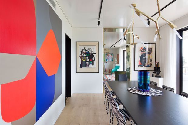 Contemporary dining area with a long dark dining table, patterned dining chairs, sculptural brass pendant lighting, light timber flooring, mirrored wall panels, and bold abstract artwork in vivid red, blue, grey, and orange. The space opens toward bright glazing on the right and includes colourful artwork and furnishings beyond the dining zone.