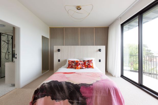 Bright contemporary bedroom with a double bed positioned against a full-height timber-look built-in headboard and integrated bedside lights and switches. The room has pale walls, soft carpet, sheer curtains, a sculptural ceiling light, and large black-framed sliding glass doors opening to a balcony. A bathroom is partially visible through the doorway at left.