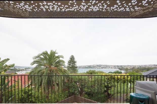 A covered outdoor terrace or balcony overlooking a leafy suburban landscape and a body of water. The space has a black metal balustrade, patterned overhead screening or pergola structure, garden planting, and outdoor seating at the right edge. The view and built elements create an airy, relaxed setting.