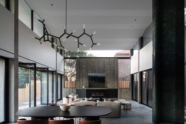 A sophisticated double-height living room with a large open-plan layout, extensive black-framed glazing, polished concrete flooring, and a dramatic branching linear pendant light. A dark timber feature wall houses the television and fireplace, with a large sectional sofa and dining table in the foreground. The space connects visually to a fenced outdoor courtyard and neighbouring brickwork.