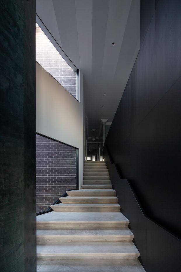 A dramatic interior stairway rises through a narrow, tall circulation space. Broad pale concrete or stone treads contrast with dark charcoal wall panelling and a dark stair balustrade. Exposed grey brick walls and high slot-like glazing bring in subdued natural light, while the ceiling has recessed downlights and linear panel detailing.