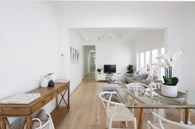 Bright, open-plan residential interior combining a dining area and living room. The space features white walls and ceiling, light timber flooring, large white-framed windows, a glass dining table with white woven chairs, a neutral sofa, a media console, indoor plants, and a timber console table. A wide opening connects the living area to a hallway or adjacent room.