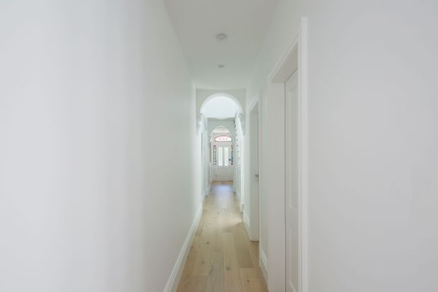 A narrow, bright residential hallway with white walls, white trim and doors, light timber flooring, recessed ceiling lights, and a decorative arched opening leading toward a glazed entry door.