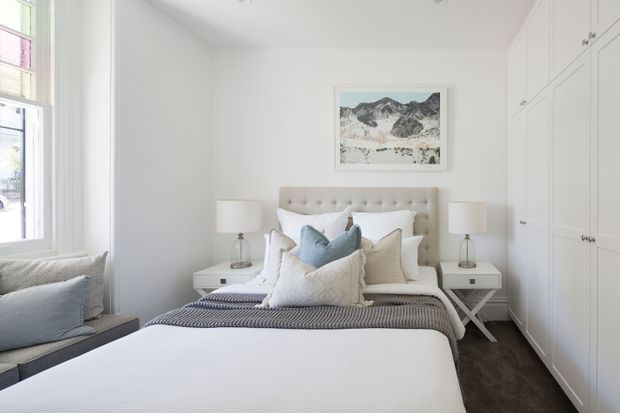 Bright, refined bedroom featuring a large upholstered button-tufted headboard, layered white and pale blue bedding, matching bedside tables and lamps, a built-in wall of white wardrobe cabinetry, a window seat, and dark carpeting. A framed mountain artwork provides a subtle focal point.