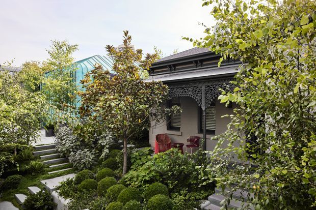 A landscaped courtyard garden surrounding a compact heritage-style dwelling or pavilion. The scene features clipped rounded shrubs, layered planting, stone steps and paths, a shaded verandah with ornate dark fretwork and columns, and a modern turquoise glazed structure visible behind the garden. Outdoor seating sits beneath the verandah.