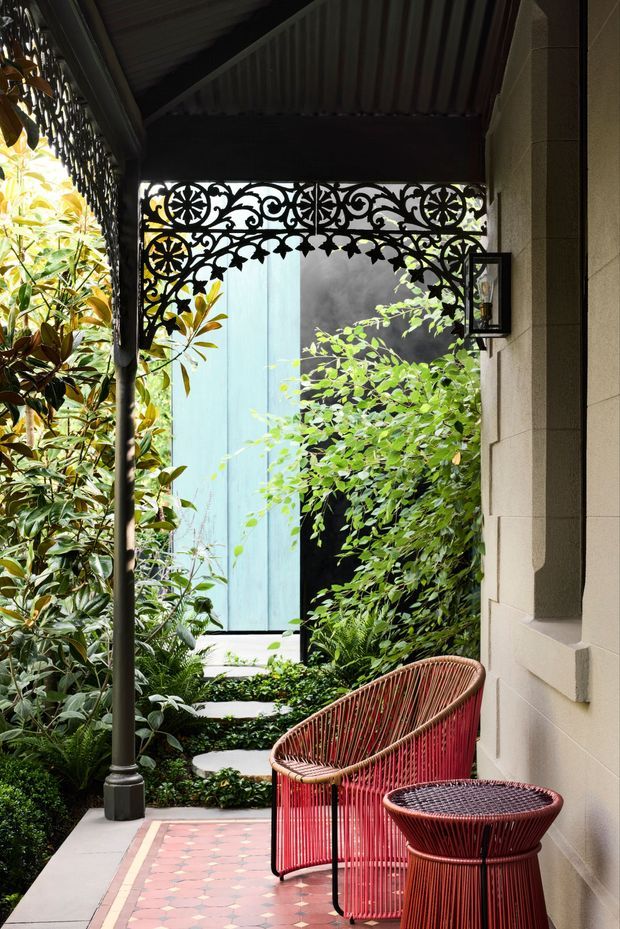 A lush, shaded outdoor verandah or terrace beside a rendered masonry wall, featuring ornate black decorative metal screening beneath a corrugated roof, a classic lantern, patterned terracotta-and-cream tiled flooring, and red woven outdoor lounge chairs with a matching side table. Dense leafy planting surrounds a stepping-stone path and a pale blue exterior wall or door is visible beyond.