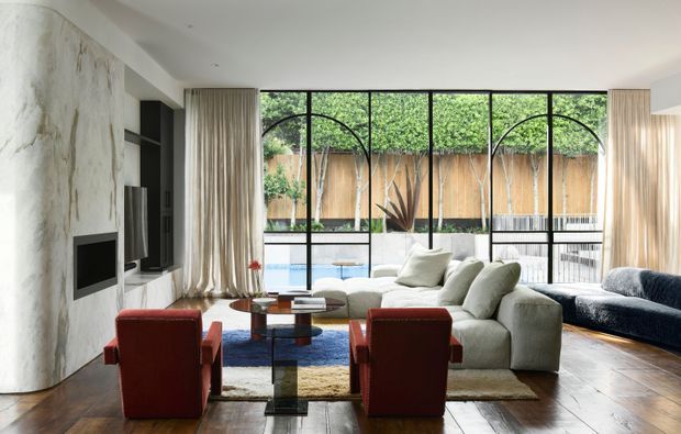 A sophisticated contemporary living room with a large sectional sofa, red lounge chairs, a dark blue sofa, and a low glass-and-metal coffee table. The room features a dramatic pale marble-look feature wall with built-in black media joinery, full-height black-framed glazing with arched detailing, and curtains opening to a private courtyard or pool area. Timber flooring and layered rugs add warmth to the otherwise refined neutral scheme.