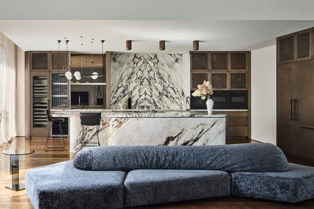 A refined open-plan kitchen and living area viewed from behind a large dark blue-grey sectional sofa. The kitchen features dark timber cabinetry, a prominent veined marble island and matching full-height splashback, integrated appliances, pendant lighting, and a breakfast bar. Light curtains and timber flooring soften the sophisticated, high-contrast interior.