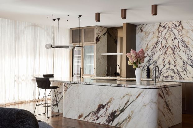 A refined open-plan kitchen dominated by a large curved island clad in dramatic veined natural stone, with matching stone splashback and cabinetry accents. Dark timber cabinetry, a black sink tap, timber flooring, sheer floor-to-ceiling curtains, sculptural pendant lighting, and a pair of bar stools create a luxurious, warm contemporary interior.