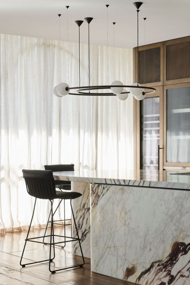 A refined open-plan kitchen and dining area with a large veined marble waterfall island, dark timber built-in cabinetry, sheer full-height curtains, black bar stools, and a sculptural ring pendant light with globe accents. The space is bright and softly filtered by daylight, with warm timber flooring and a restrained neutral palette.