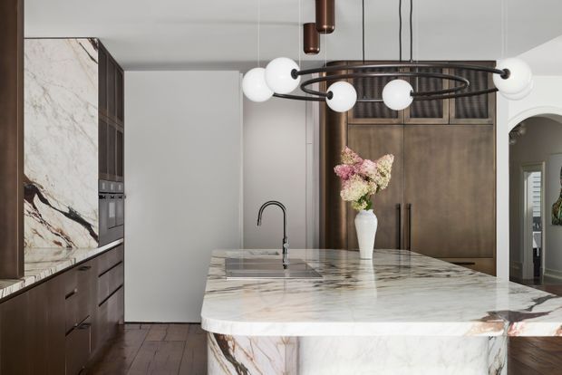 A sophisticated contemporary kitchen centered on a large waterfall island clad in heavily veined white marble. Dark timber cabinetry lines the left wall and continues behind the island, with integrated appliances and a concealed or minimal splashback. A black gooseneck tap and undermount sink sit within the island, while a sculptural black circular pendant with globe lights hangs above. An arched doorway and glimpses of adjoining rooms appear at right.