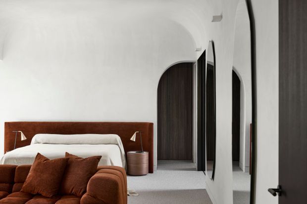 A refined bedroom interior with a warm rust-brown upholstered bedhead and matching plush seating in the foreground. White bedding, pale grey carpet, crisp white walls, arched doorways, dark timber doors, and slim wall-mounted bedside lights create a calm, modern composition. Full-height mirrored wardrobe doors reflect the room and reinforce the sense of depth.