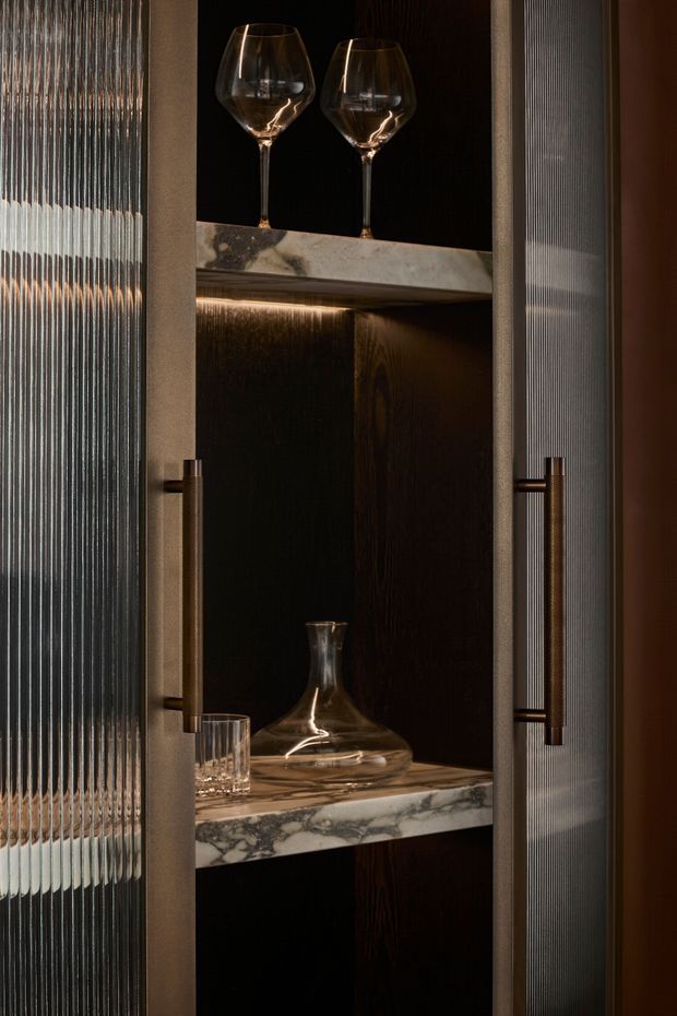 Close-up of a dark, luxurious built-in bar or display cabinet with ribbed translucent glass doors, bronze-toned vertical handles, dark timber interior, dramatic integrated lighting, and veined marble shelves. Crystal wine glasses, a decanter, and tumblers are displayed inside.