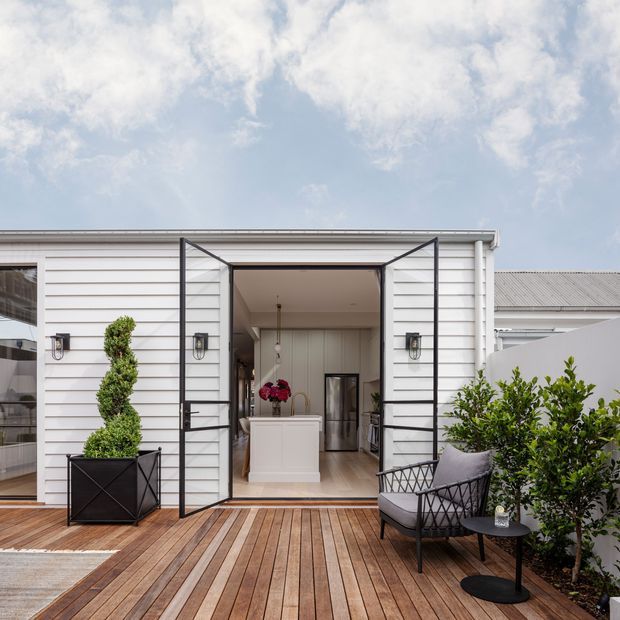 A bright exterior view of a compact white weatherboard-clad building opening onto a timber deck. Black-framed glazed folding doors reveal a light-filled interior with a central white island or counter, while potted topiary, planting, and an outdoor lounge chair furnish the deck. The simple low-pitched roof and clean-lined detailing create a contemporary coastal feel.