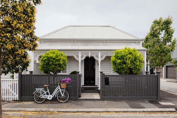 Front exterior view of a traditional single-storey weatherboard house with a low-pitched hipped metal roof, ornate decorative fretwork verandah, black entrance door, and enclosed front garden. The property has a charcoal vertical-slat fence, clipped shrubs, a white picket side fence, and a bicycle at the street edge.