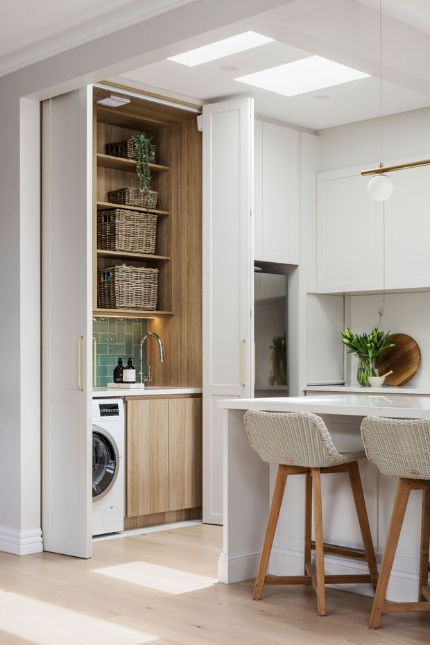 Bright, refined transitional kitchen with a concealed laundry area behind tall paneled doors. The space features white shaker-style cabinetry, a pale stone island, light timber flooring, a warm timber-lined utility cupboard with open shelving and woven baskets, and a muted green tiled splashback. Two woven bar stools sit at the island, while integrated lighting and brass accents add detail.