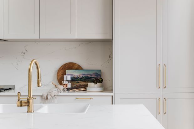 Bright, refined kitchen interior with pale grey built-in cabinetry, a white marble-look splashback and benchtops, an undermount sink, and a prominent brushed brass gooseneck tap. Tall pantry-style cabinetry with brass handles lines the right side, while a small styled vignette sits on the rear counter.