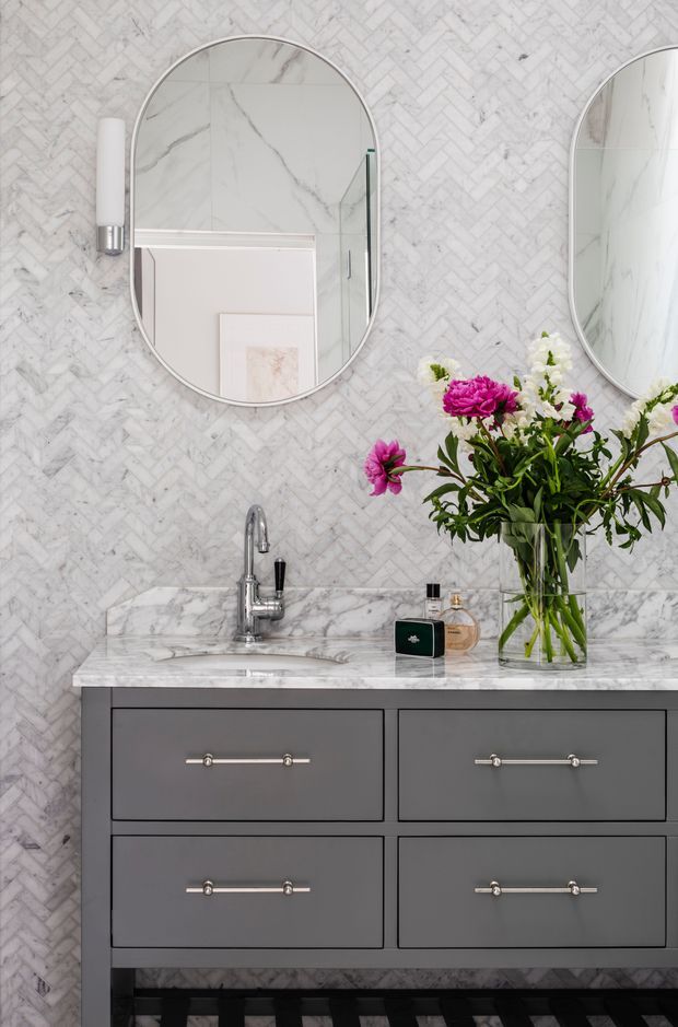 A refined bathroom vanity featuring a grey four-drawer floating-style cabinet with polished metal handles, a white marble countertop and integrated basin, chrome tapware, and a full-height white-and-grey marble herringbone wall finish. Two rounded-arched mirrors and contemporary wall sconces sit above the vanity, with a clear glass vase of pink and white flowers adding colour.