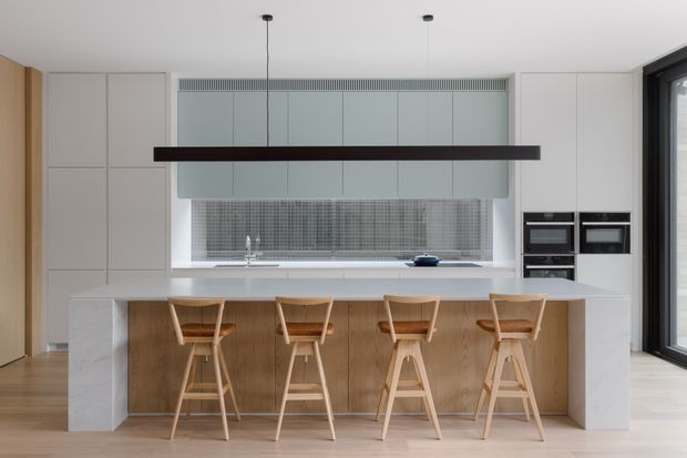 A bright, refined contemporary kitchen featuring a large white stone island with integrated seating for four, pale wood cabinetry and stools, full-height light grey cabinetry, a grey mosaic tile splashback, and built-in black appliances. A long dark linear pendant hangs above the island, with timber flooring and a large glazed opening at the side.