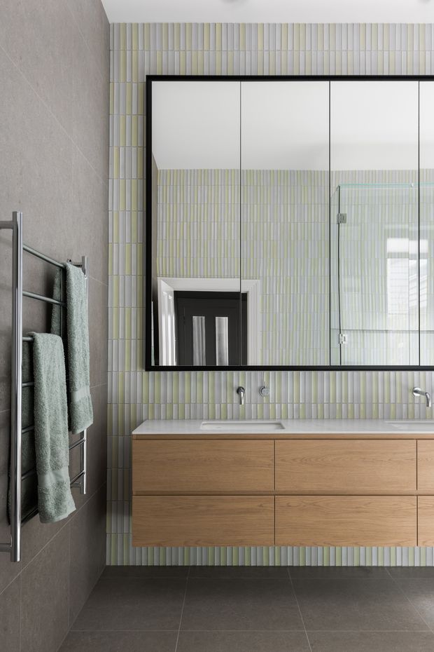 Contemporary bathroom vanity with a floating warm timber double cabinet, pale stone countertop, wall-mounted basins and brushed metal taps. The walls are clad in narrow vertically arranged pale green, white and grey tiles, contrasted by a large black-framed mirrored cabinet. A dark grey tiled floor and matching grey wall create a refined, modern scheme, with a chrome heated towel rail and muted green towels at left.