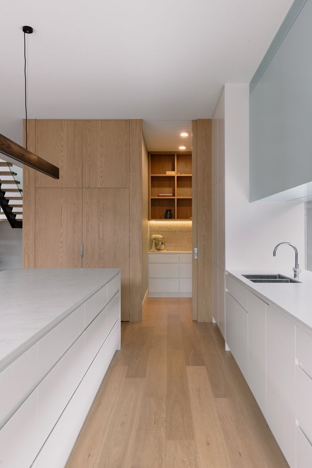 A bright, contemporary galley-style kitchen with a large white island, white built-in cabinetry, pale timber wall cabinetry and flooring, and a compact integrated butler's pantry or appliance nook at the rear. A sink with a curved chrome tap sits along the right-hand benchtop, while open timber shelving provides a warm feature. A floating stair edge and linear pendant light are visible at the left.