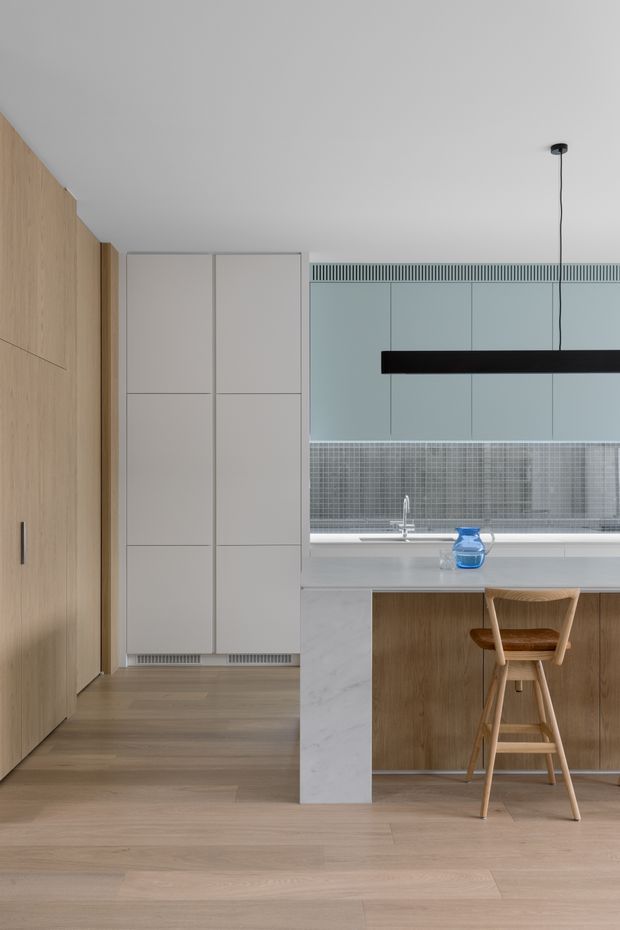 A refined contemporary kitchen with a warm timber veneer wall and concealed cabinetry on the left, pale blue-grey upper cabinetry, and a narrow grey mosaic tile splashback. The island features a white marble-look benchtop, timber cabinetry, and a single timber bar stool. Light timber flooring, white ceilings, integrated ventilation detailing, and a slim black linear pendant create a clean, understated interior.
