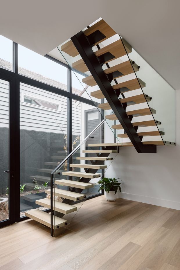 A bright contemporary stairway with floating light timber treads supported by a dark central steel spine and enclosed by clear glass balustrades. The stair rises beside full-height black-framed glazing overlooking a small internal courtyard, with pale timber flooring, white walls, and a potted plant. The design is open, minimalist, and highly detailed.