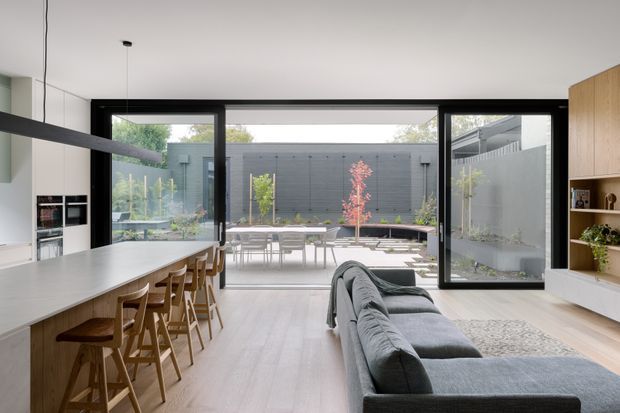 Open-plan contemporary kitchen, dining and living area opening through large black-framed glazed doors to a landscaped courtyard. The room features a long pale island with timber bar stools, integrated cabinetry and appliances, a grey sectional sofa, light timber flooring, built-in timber shelving, and a minimalist linear pendant light.
