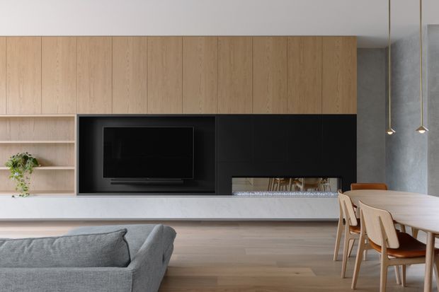 A refined open-plan living and dining area with a built-in timber and matte-black media wall. The room features a wall-mounted television, low linear fireplace, pale stone or concrete-look hearth, light timber flooring, a grey upholstered sofa, and a timber dining table with matching chairs. Minimal pendant lights and restrained styling create a calm, contemporary interior.