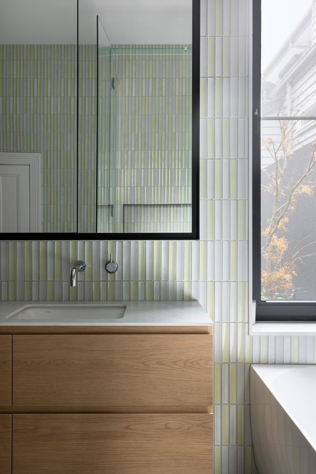 Contemporary bathroom vanity with a wall-mounted timber drawer cabinet, integrated pale benchtop and rectangular basin, chrome wall-mounted tapware, and a large black-framed mirror. The walls are clad in narrow vertical glazed tiles in muted sage green, pale green, cream and grey tones. A black-framed window brings in natural light beside the vanity.