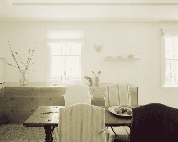 A softly lit, monochromatic dining room with a dark wooden dining table and chairs, upholstered slipcovered seating, a long low built-in sideboard beneath the windows, and a simple floating shelf with understated decor. Tall divided-light windows, pale walls, and restrained furnishings create a calm, traditional interior.