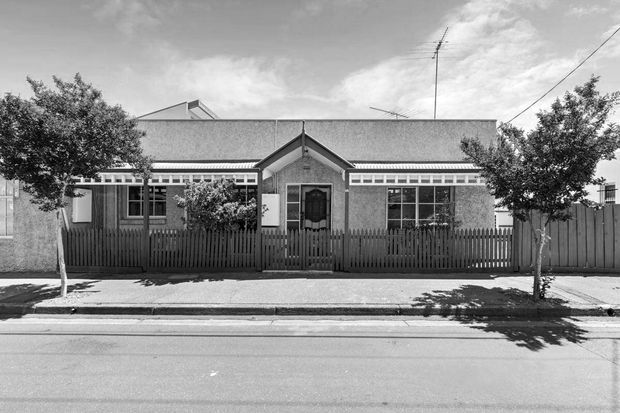 Black-and-white street-facing view of a single-storey residential building with a symmetrical gabled entrance porch, broad pitched roof, textured rendered walls, multi-pane windows, decorative clerestory-style glazing, and a timber picket fence enclosing the front garden. Mature street trees and a paved footpath frame the facade.