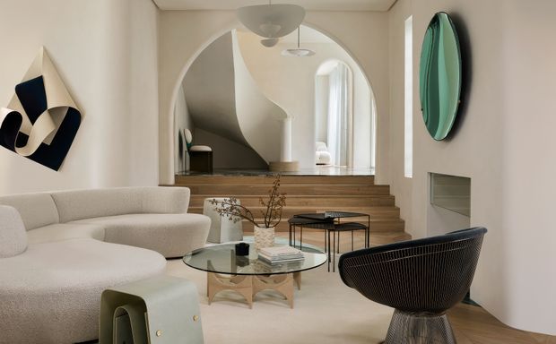 A refined contemporary living room with a curved cream sofa, sculptural lounge chair, glass coffee table, and abstract wall art. Warm timber steps lead through a large arched opening toward a softly lit upper-level area, with built-in wall niches and rounded architectural forms throughout.