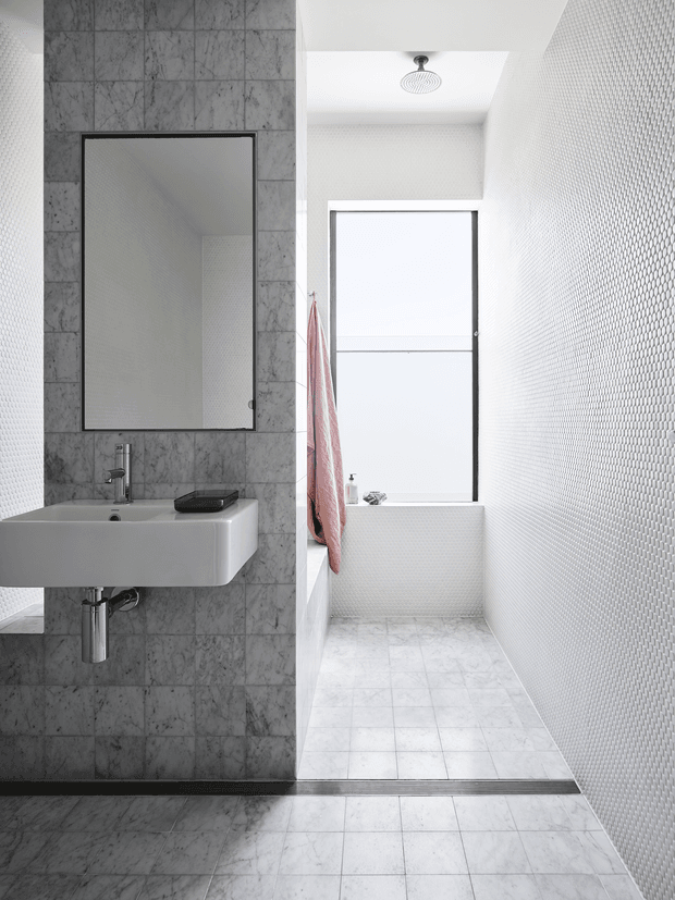 Bright contemporary bathroom with a wall-mounted white basin and exposed chrome plumbing, large black-framed mirror, grey marble-look tiled feature wall, white small-format tiled walls, and pale stone-look floor tiles. A walk-in shower area with ceiling-mounted shower head is positioned beyond the basin, with a frosted window and pink towel adding a subtle accent.