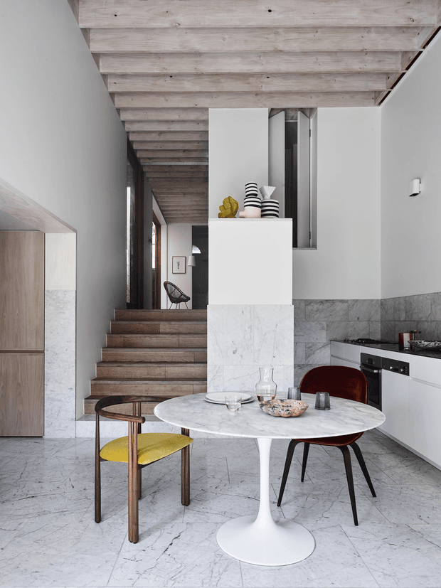 A refined open-plan kitchen and dining area with a round white pedestal table, sculptural dining chairs, white cabinetry, integrated appliances, and a marble-clad island or partition. A timber-lined ceiling continues over a short flight of timber stairs leading to a hallway or upper level. The space combines pale stone surfaces, white walls, warm timber, and restrained decorative objects in a bright, sophisticated interior.