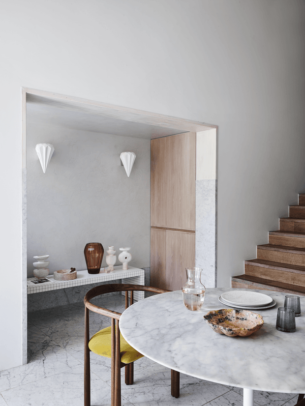 A refined open-plan dining area with a round white marble dining table, sculptural wooden chairs with yellow upholstery, and a built-in display niche. The niche has a textured pale plaster wall, white tiled bench, light timber cabinetry, and decorative ceramic and glass objects. A timber staircase rises at the right, while white and grey marble-like flooring and soft grey walls create a calm, contemporary interior.