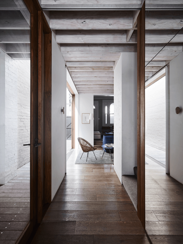 A long, bright residential hallway viewed through a dark timber-framed opening, with white-painted masonry walls, exposed pale timber ceiling beams, and wide timber floorboards. The corridor leads to a small living area furnished with a woven lounge chair, low table, blue sofa, and rug. Full-height glazing and reflective surfaces create layered views along the passage, while a carpeted stair or seating edge is visible to the right.
