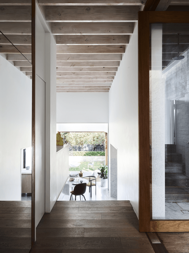 A tall, minimalist interior corridor or landing with white walls, dark timber flooring, and exposed pale timber ceiling joists. The passage overlooks a bright dining area and garden through a large opening, while a wood-framed glazed opening on the right reveals a dark tiled bathroom or wet area. The composition emphasizes strong vertical lines, natural light, and a contrast between light plaster surfaces and warm timber elements.