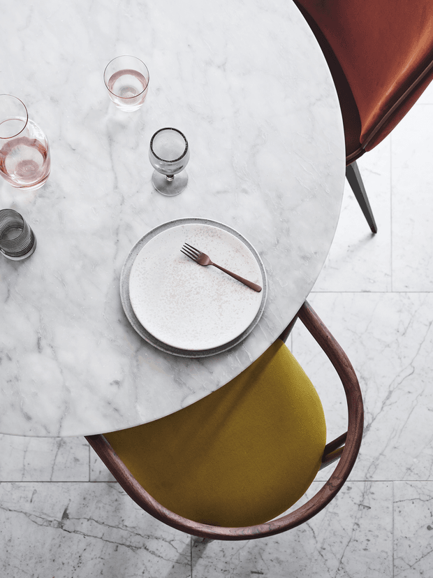 Top-down view of a refined dining setting: a round white marble table with subtle veining, ceramic plates, a fork, assorted drinking glasses, and a sculptural dining chair with a mustard upholstered seat and dark timber curved frame. The table sits over a pale veined stone or marble floor, creating a sophisticated, warm contemporary composition.