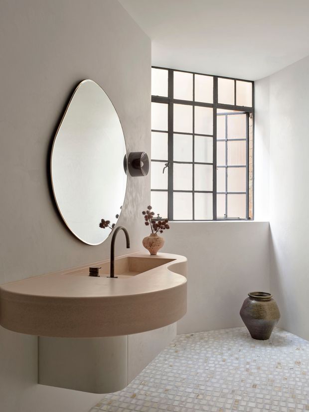 A refined minimalist bathroom featuring a sculptural curved integrated vanity with a pale stone or plaster-like finish, a matching curved basin, dark tapware, and a large round wall mirror. The room has white walls, a black-framed industrial-style grid window, and a small-format pale mosaic tile floor. A ceramic vessel provides a subtle decorative accent.
