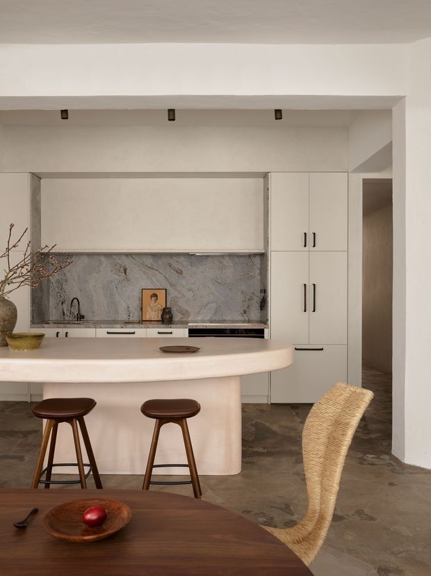Warm, sculptural kitchen and dining area with a curved pale island, integrated cabinetry, stone backsplash, dark stone floor, and timber bar stools. A dark timber dining table with a woven chair occupies the foreground. The kitchen features restrained neutral finishes, black hardware, concealed appliances, and softly rounded forms.