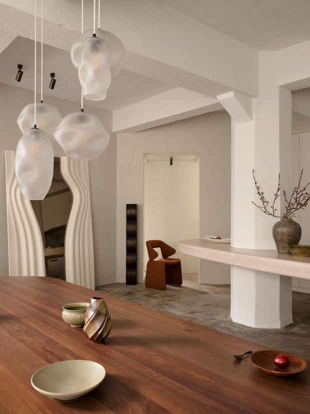 Warm, sculptural contemporary dining and living interior with a large dark timber dining table in the foreground, softly plastered cream walls, chunky structural columns and beams, a curved built-in breakfast counter, and distinctive organic pendant lights. A wavy-framed mirror, rust-coloured lounge chair, ceramic vessels, and an exposed textured floor add artistic character.