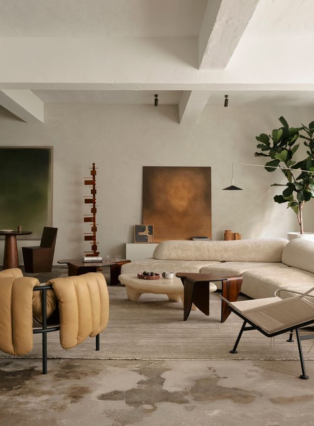 A warm, refined living room with a large cream modular sectional sofa, sculptural upholstered armchairs, dark wood and metal coffee tables, and a textured neutral rug over a polished concrete floor. The room features white plaster walls, exposed painted structural ceiling beams, minimalist pendant lighting, abstract artwork, and a tall indoor tree. The overall look blends contemporary and modern classic furnishings with a calm, earthy palette.