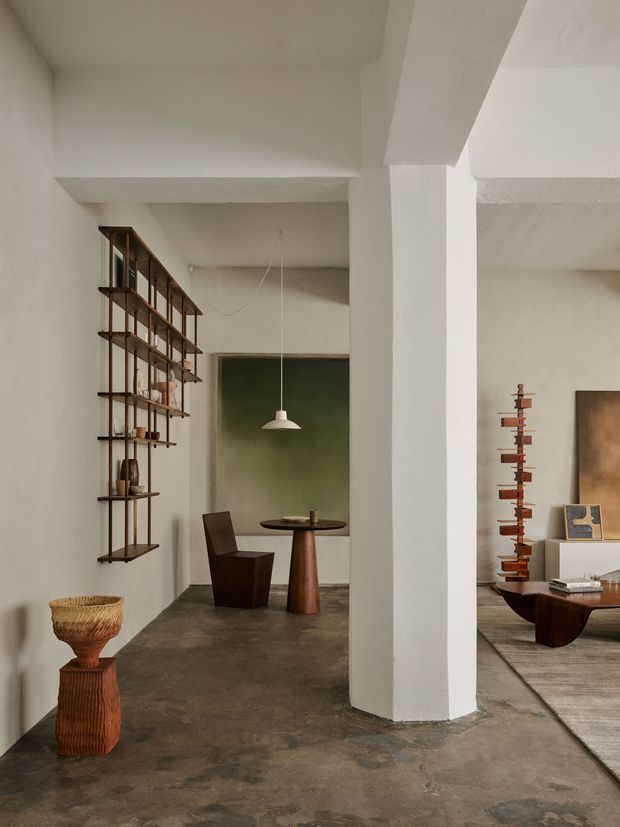 A warm, minimalist interior with a concrete floor, white plastered walls and exposed structural columns. A narrow dining area sits beyond the central column, furnished with a small dark wood table, sculptural chairs and a simple pendant light. Open wood shelving displays ceramics along the left wall, while part of a refined living area with a low coffee table, rug and artwork is visible on the right. The space has a gallery-like, earthy and curated atmosphere.