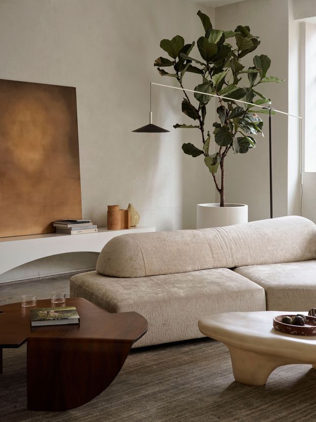 A warm, refined living room with a large curved cream sectional sofa, dark timber coffee table, sculptural light-toned side table, and a tall indoor fiddle-leaf fig in a white planter. The room features pale plaster walls, a large bronze-toned wall artwork or panel, a slim black pendant light, layered neutral textiles, and soft natural light.
